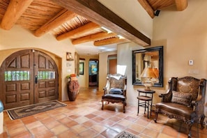 A spacious living room with a large wooden door, a fireplace, and a comfortable seating area with a chair and a sofa. - Los Valverde - Exclusive Luxury Home, Unsurpassed Views, Pool and Hot Tub! (Santa Fe)