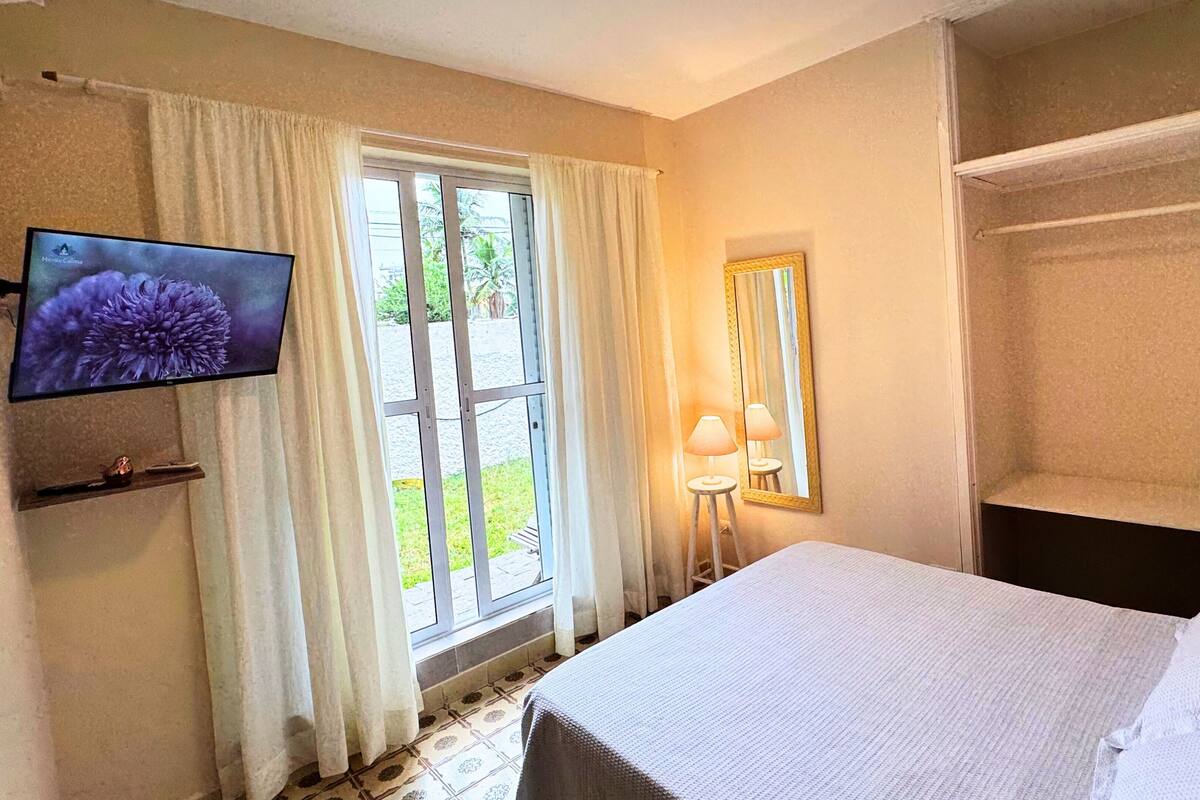 A hotel room with a bed, a television on the wall, a window with curtains, and a mirror.