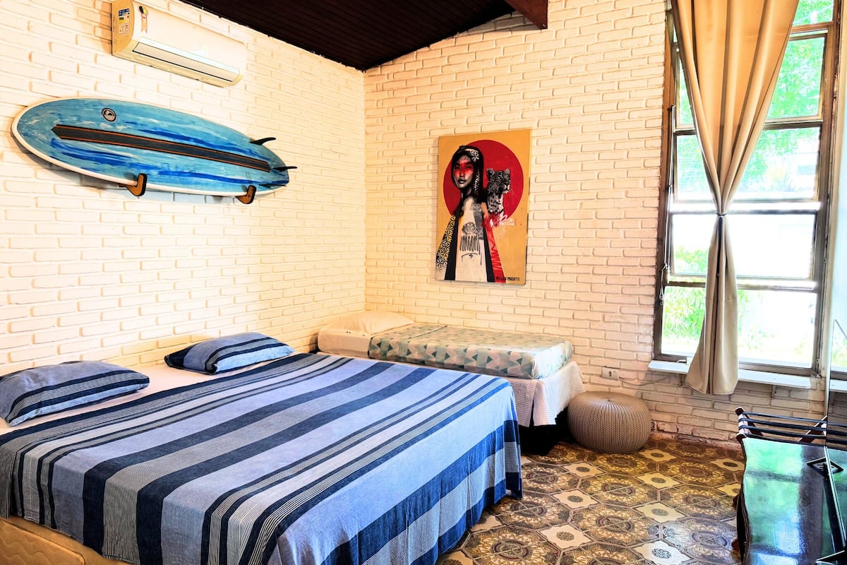 A room with two beds, a surfboard, a painting, and a window with curtains.