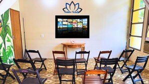 A room with a wall-mounted TV displaying a collage of images, a wooden table with a pink object, and six foldable chairs. - Tombo Beach (Guarujá)