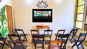 A room with a wall-mounted TV displaying a collage of images, a wooden table with a pink object, and six foldable chairs. - Tombo Beach (Guarujá)