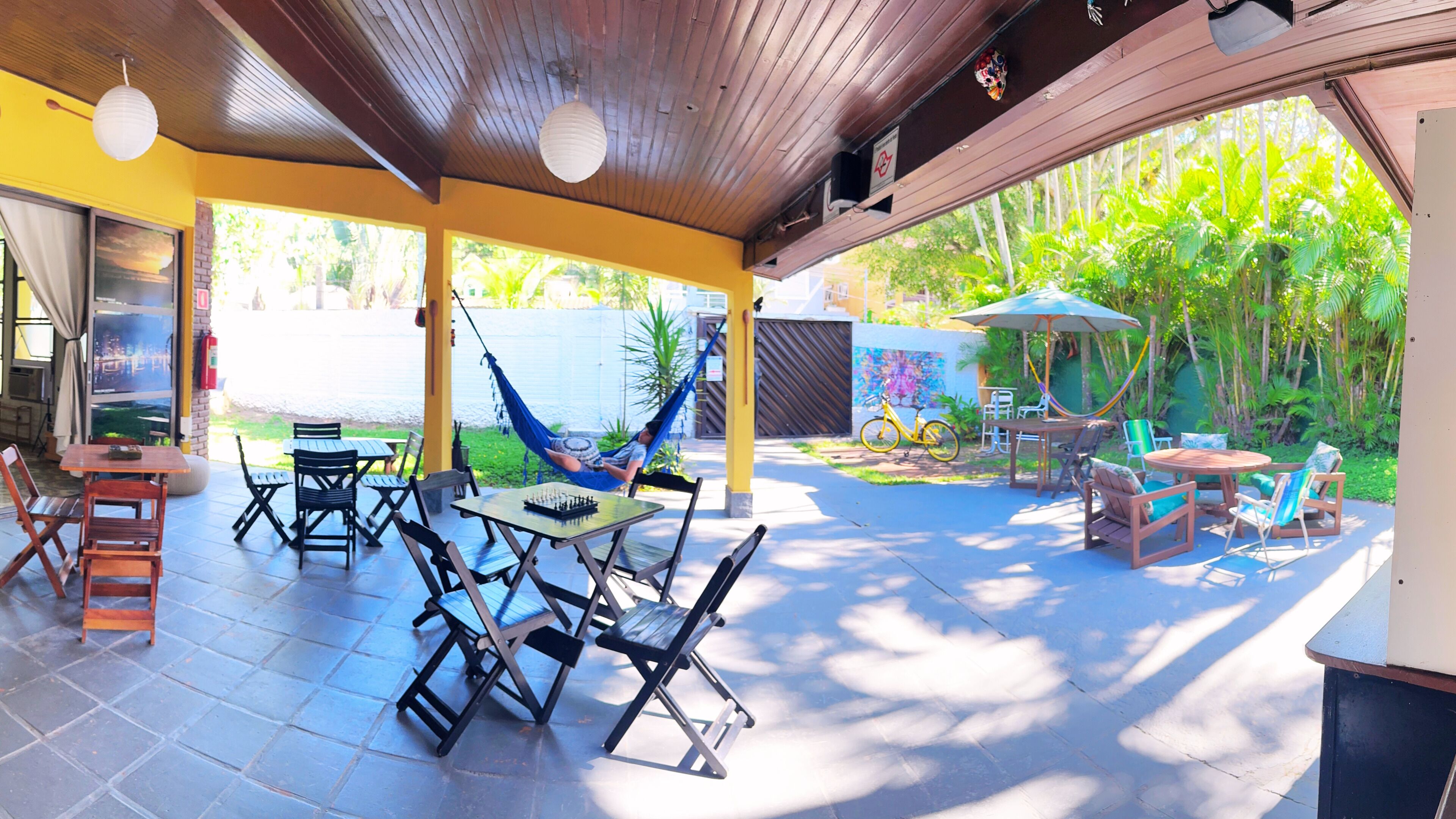A covered patio with outdoor seating, a hammock, and a view of a pool and greenery.