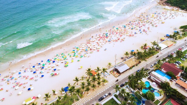 Eine Luftaufnahme eines Strandes mit zahlreichen bunten Sonnenschirmen, einer parallel zur Küste verlaufenden Straße und einem Gebäude mit rotem Dach.
