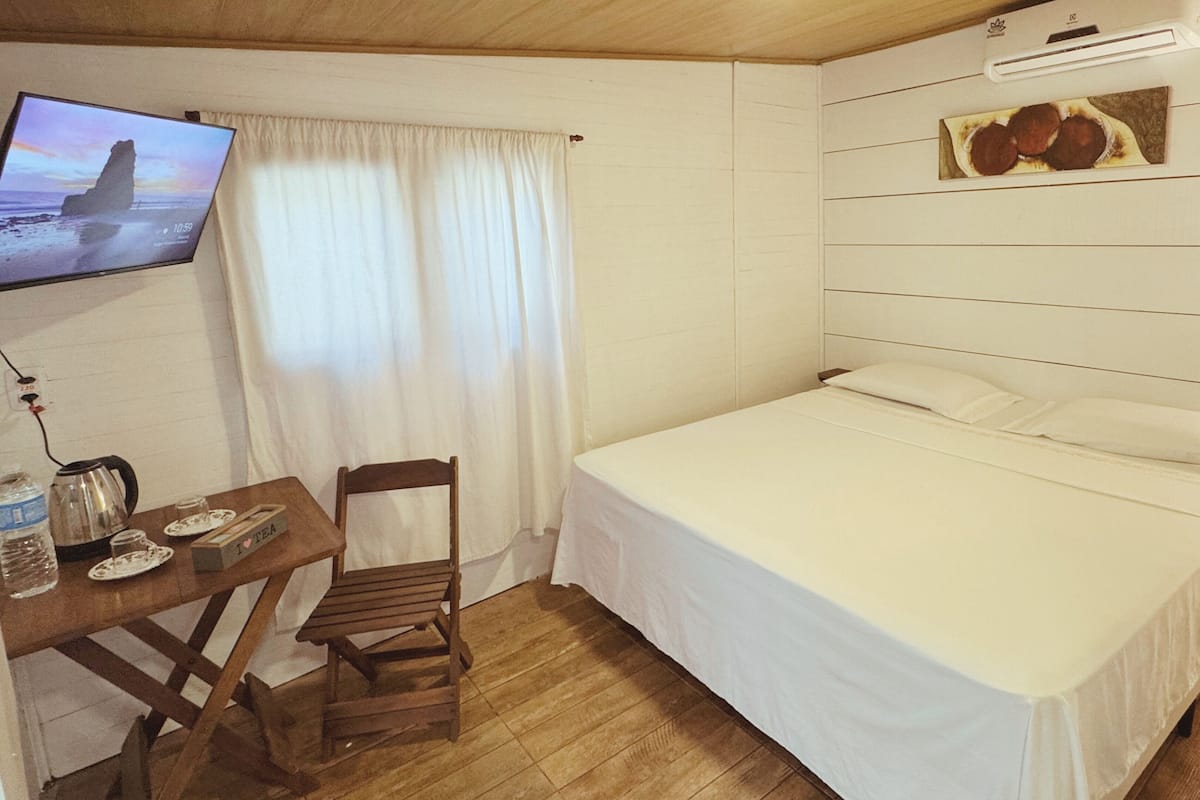 A bedroom with a bed, a desk with a kettle, a chair, and a television on the wall.