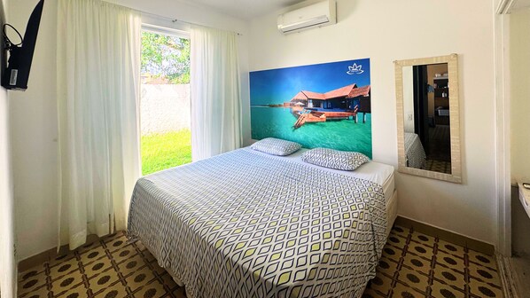 A hotel room with a bed, a window with curtains, a wall-mounted air conditioner, and a large poster of a beach scene. - Tombo Beach (Guarujá)