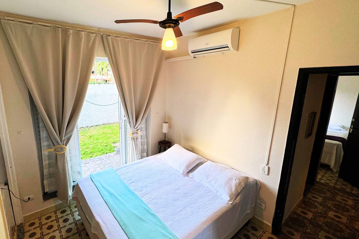 A small, neatly arranged bedroom with a bed, an air conditioning unit, a ceiling fan, and a view of the outside.