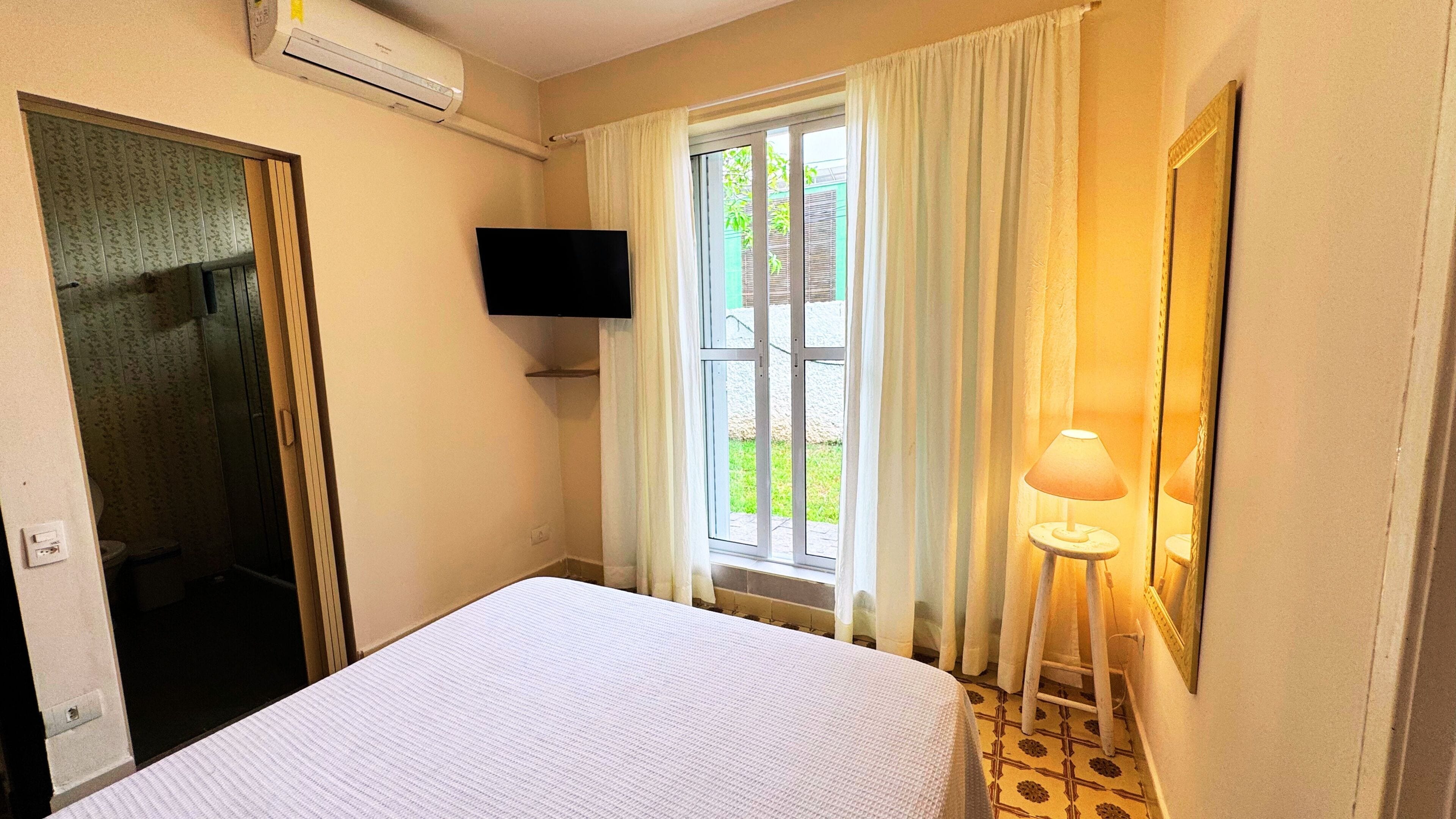A hotel room with a bed, a TV mounted on the wall, a window with curtains, a small table with a lamp, and an air conditioning unit on the wall.