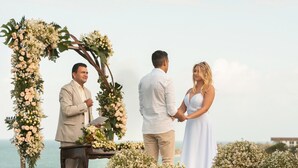 Eine Hochzeitszeremonie mit einem floralen Bogen und Gästen, die auf einer Wiese mit Blick auf das Meer stehen.