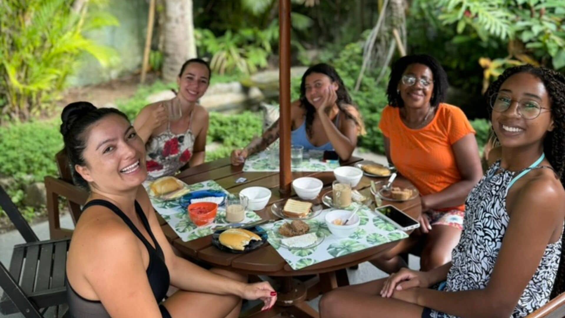 A group of people sitting around a table with food and drinks, outdoors with greenery in the background.
