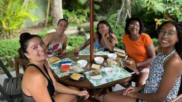 A group of people sitting around a table with food and drinks, outdoors with greenery in the background.