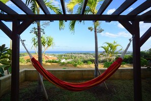 A hammock under a wooden pergola with a view of palm trees and a distant landscape. - Somos (Nakijin)