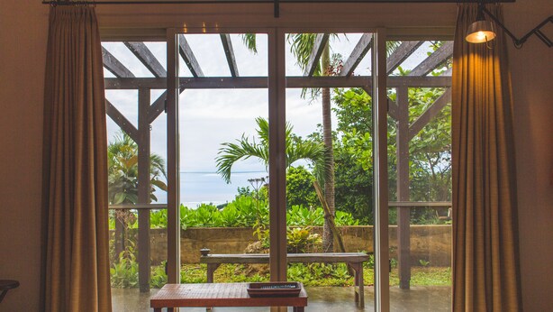 A room with a large sliding glass door leading to an outdoor area with a bench and greenery. - Somos (Nakijin)