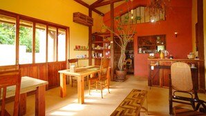 A wooden dining area with a table and chairs, a potted plant, and a kitchen area with a counter and shelves. - Somos (Nakijin)