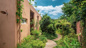 A narrow pathway leading to a building with a pink facade, surrounded by lush greenery and a clear blue sky. - Somos (Nakijin)