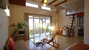 A spacious living area with a sofa, a wooden chair, a TV, a plant, and a sliding glass door leading to an outdoor area. - Somos (Nakijin)