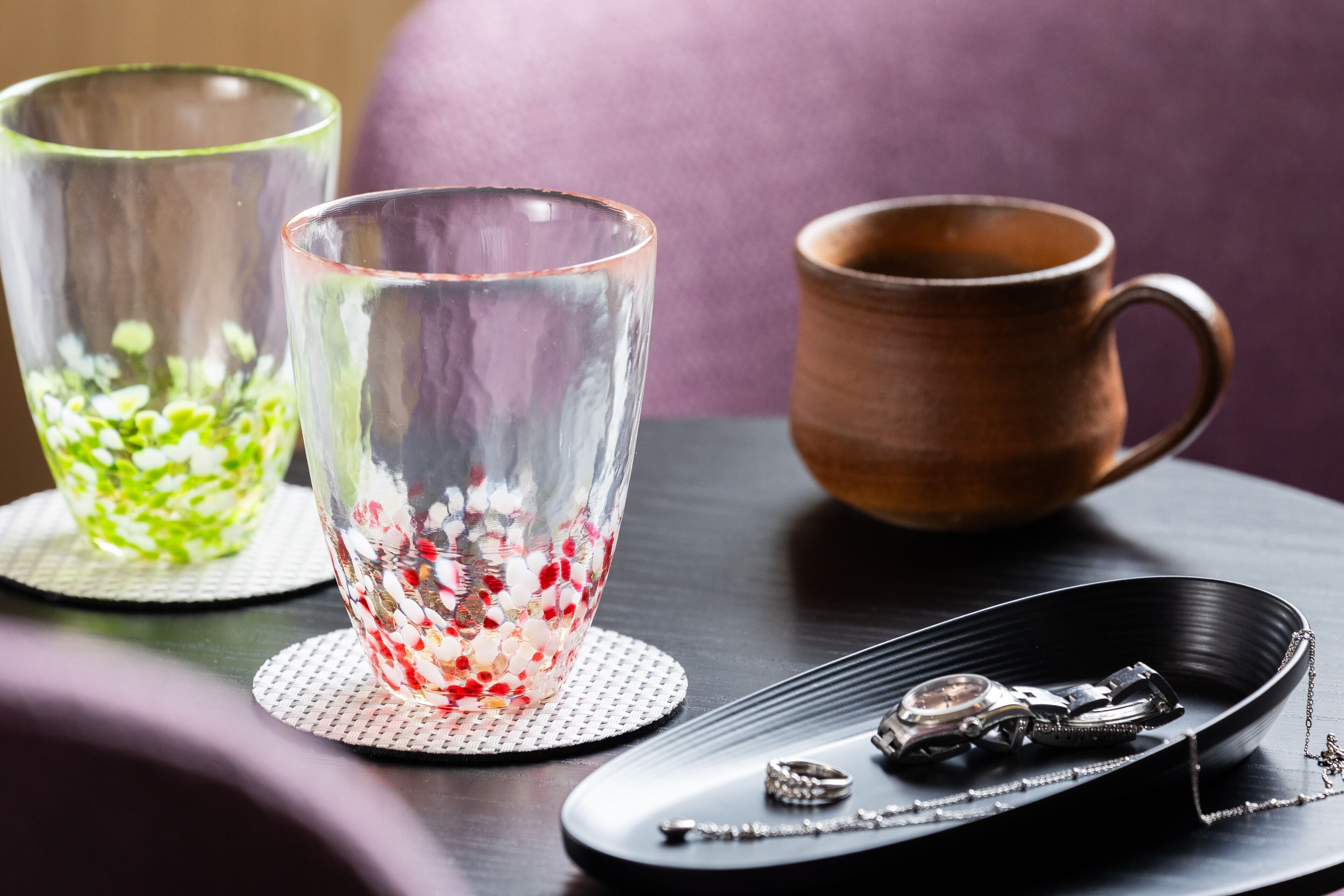Une table avec deux verres transparents, l’un contenant des perles rouges et blanches, et une tasse marron. On y trouve également une paire de boucles d’oreilles et une montre sur un plateau noir.