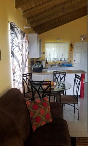 Una sala de estar acogedora con un sofá marrón y una mesa de vidrio, una cocina con armarios blancos y un ventanal, y un techo de madera.