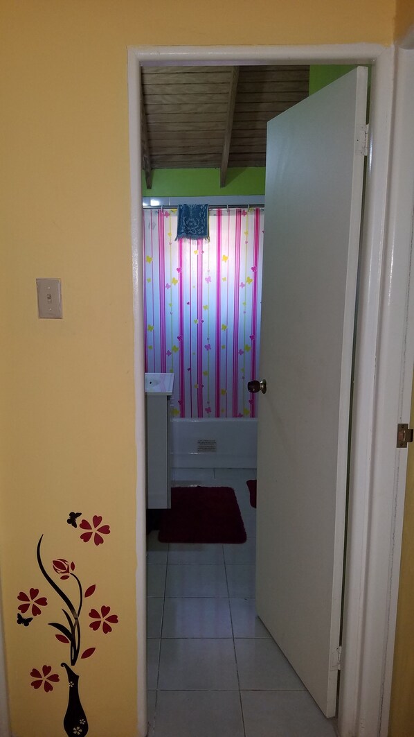 Un couloir étroit mène à une salle de bain dont les murs sont recouverts d’un papier peint à rayures, qui contient un lavabo et un tapis rouge.