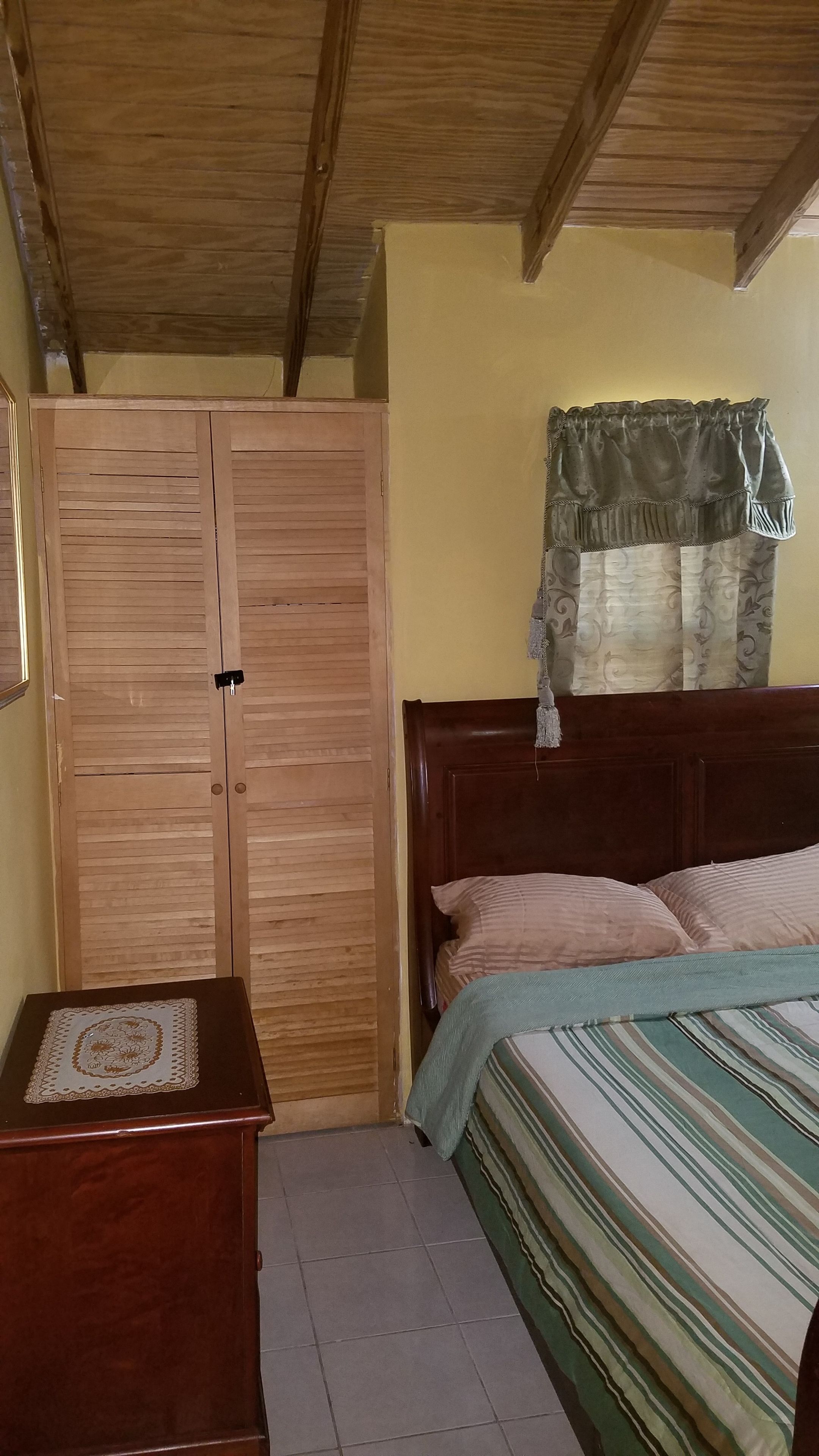Un dormitorio con un armario de madera, una mesita de noche, una cama con ropa de cama a rayas y una ventana con cortina.