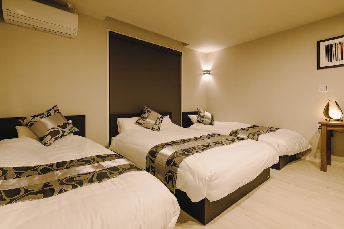 A hotel room with three beds, each with patterned bedding and a headboard. There is a bedside table with a lamp, a wall-mounted air conditioner, and a framed picture on the wall.