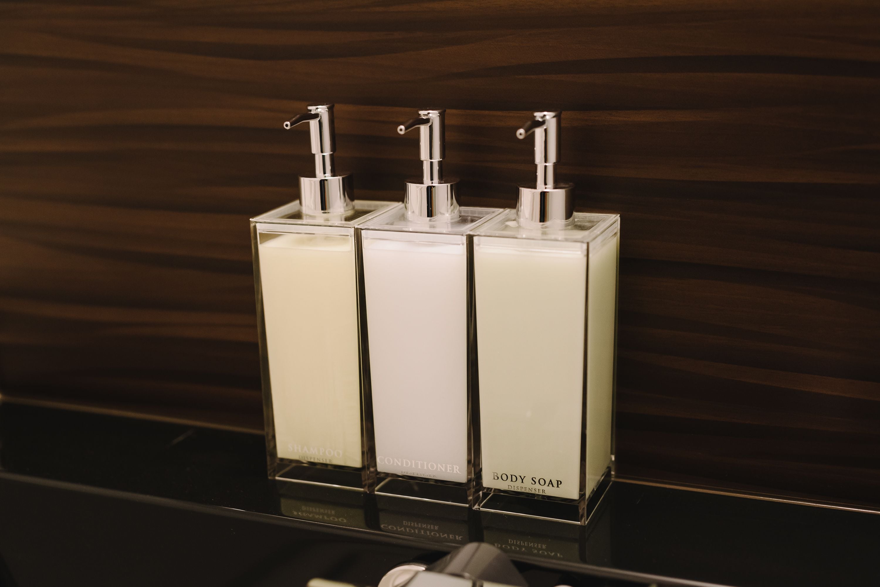 Tres dispensadores transparentes en línea, etiquetados como jabón corporal, acondicionador y champú.