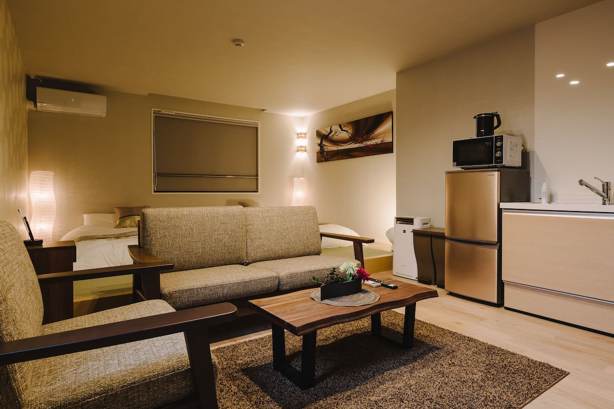 A modern living room with a sofa, coffee table, and a kitchen area with a refrigerator and microwave.