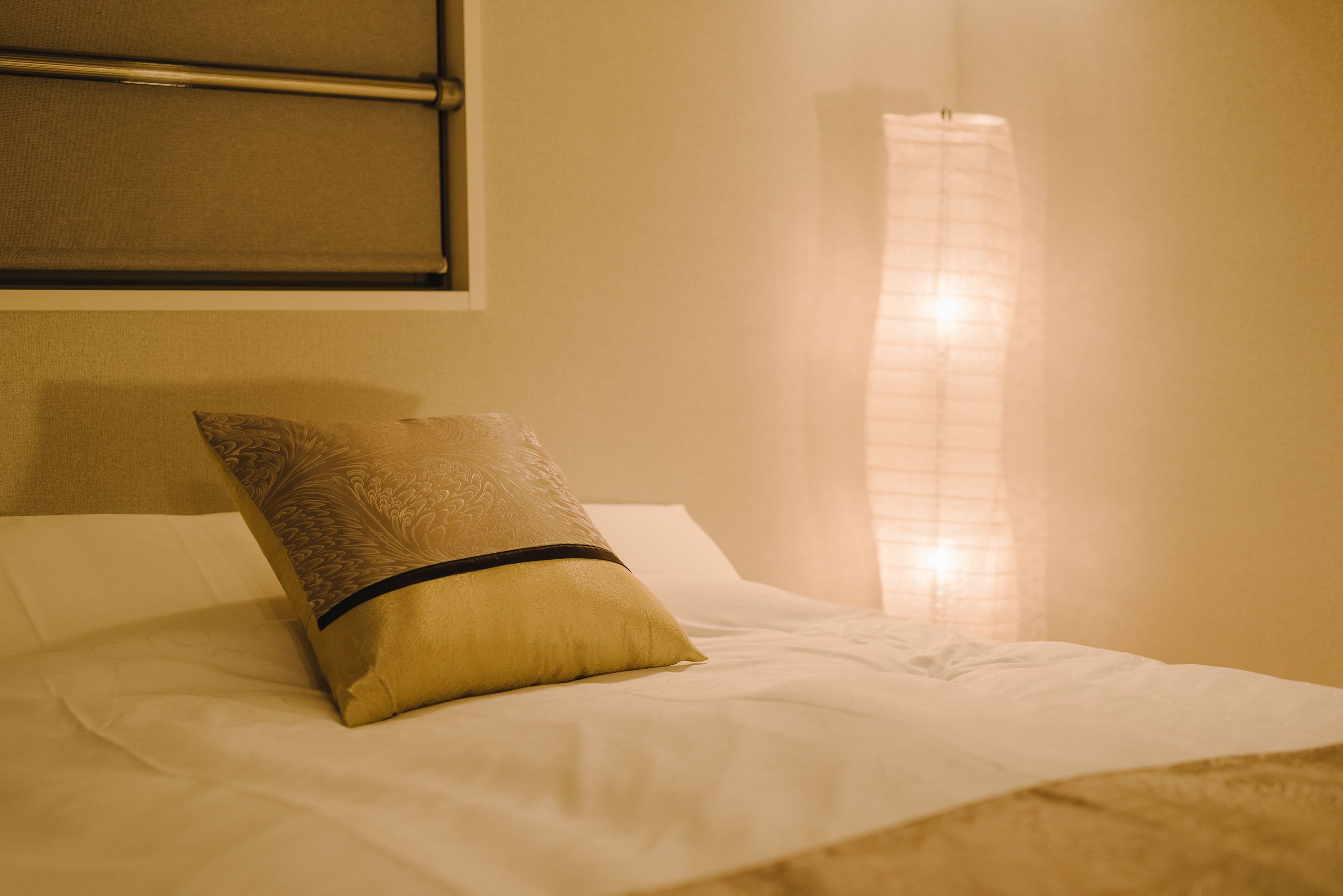 Una cama bien hecha, con un cojín estampado y uno liso, un cabecero y una ventana con persianas.