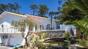 A modern house with a stone staircase leading to a balcony, surrounded by a garden with a pond and various plants.