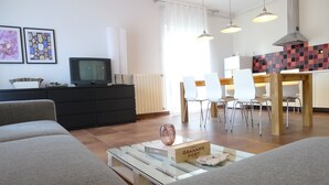 Un salon moderne avec un canapé, une table basse sur laquelle se trouvent un livre et un verre, un téléviseur posé sur un meuble noir, et un coin repas avec des chaises et une table.