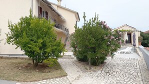 Una casa de dos pisos con balcón, un patio con baldosas y un pequeño jardín con árboles y arbustos.
