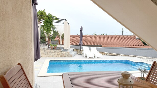Une table en bois, sur laquelle sont posés un lanternon blanc et une petite assiette, offre une vue sur une piscine bordée de chaises longues blanches et agrémentée de plantes en pot.