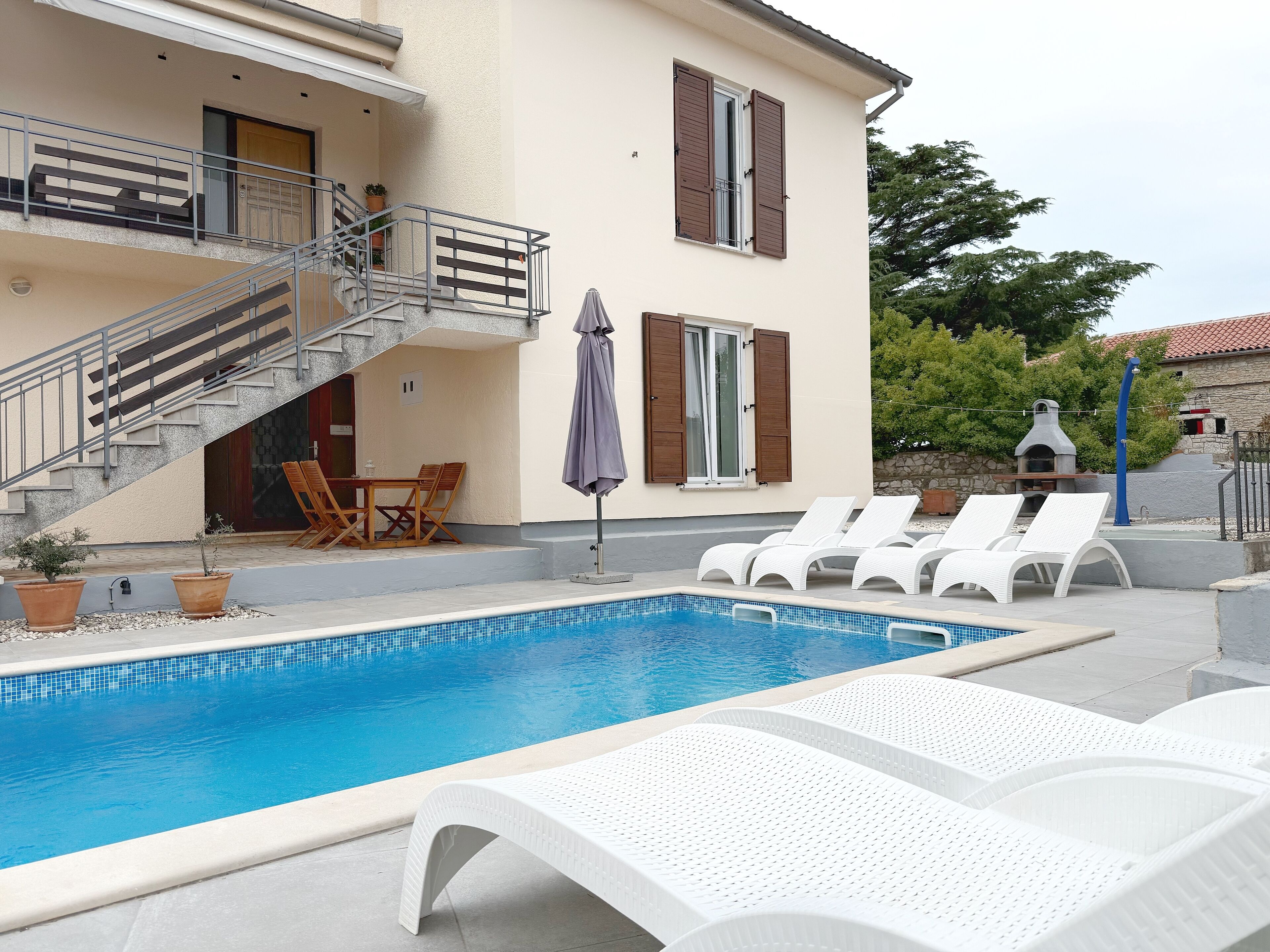 Une piscine entourée de chaises longues blanches, un escalier menant à un balcon et un espace patio avec une table et des chaises.