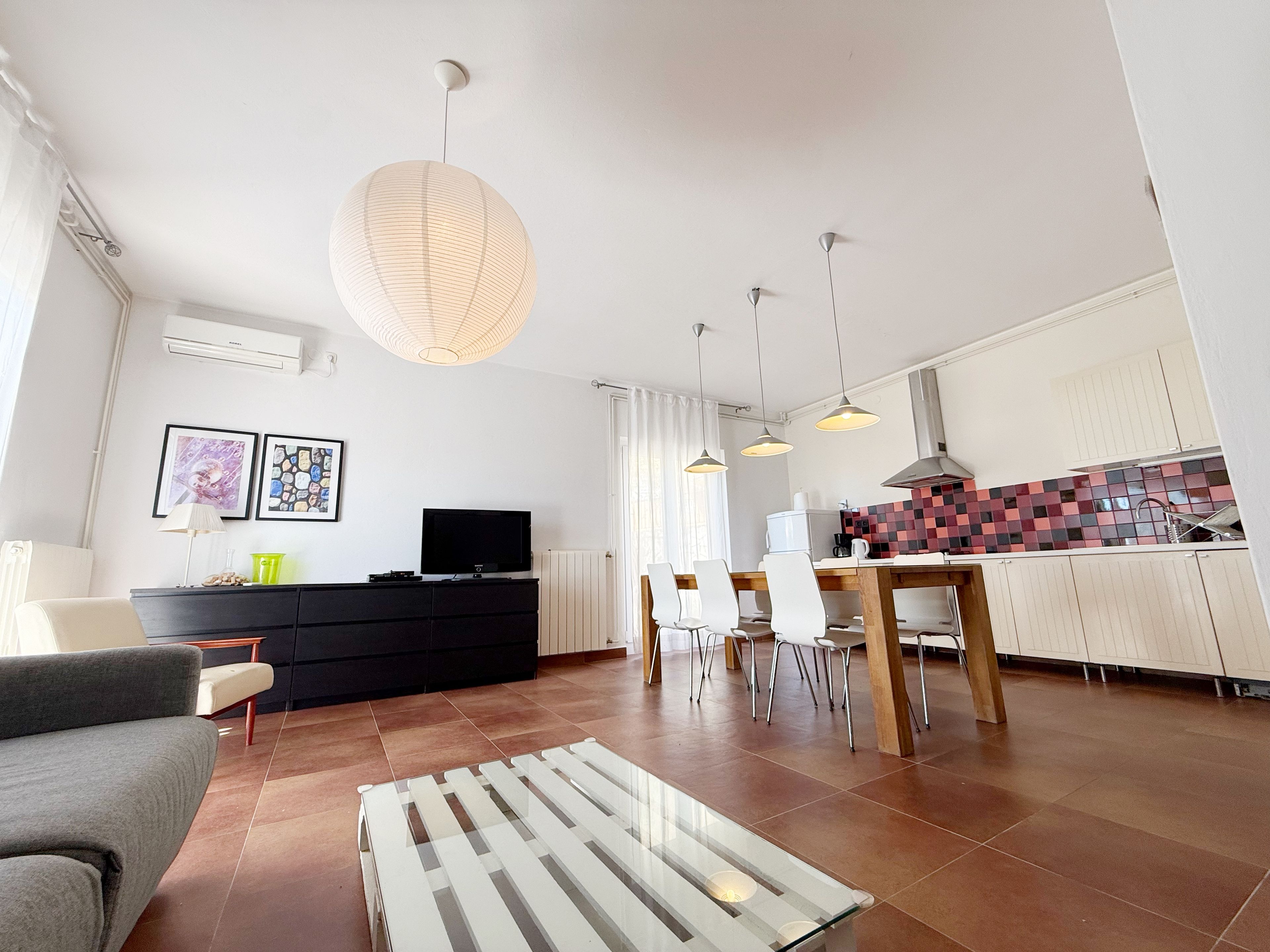 A modern living room with a sofa, a TV, a dining table with chairs, and a kitchen area with cabinets and a tiled backsplash.