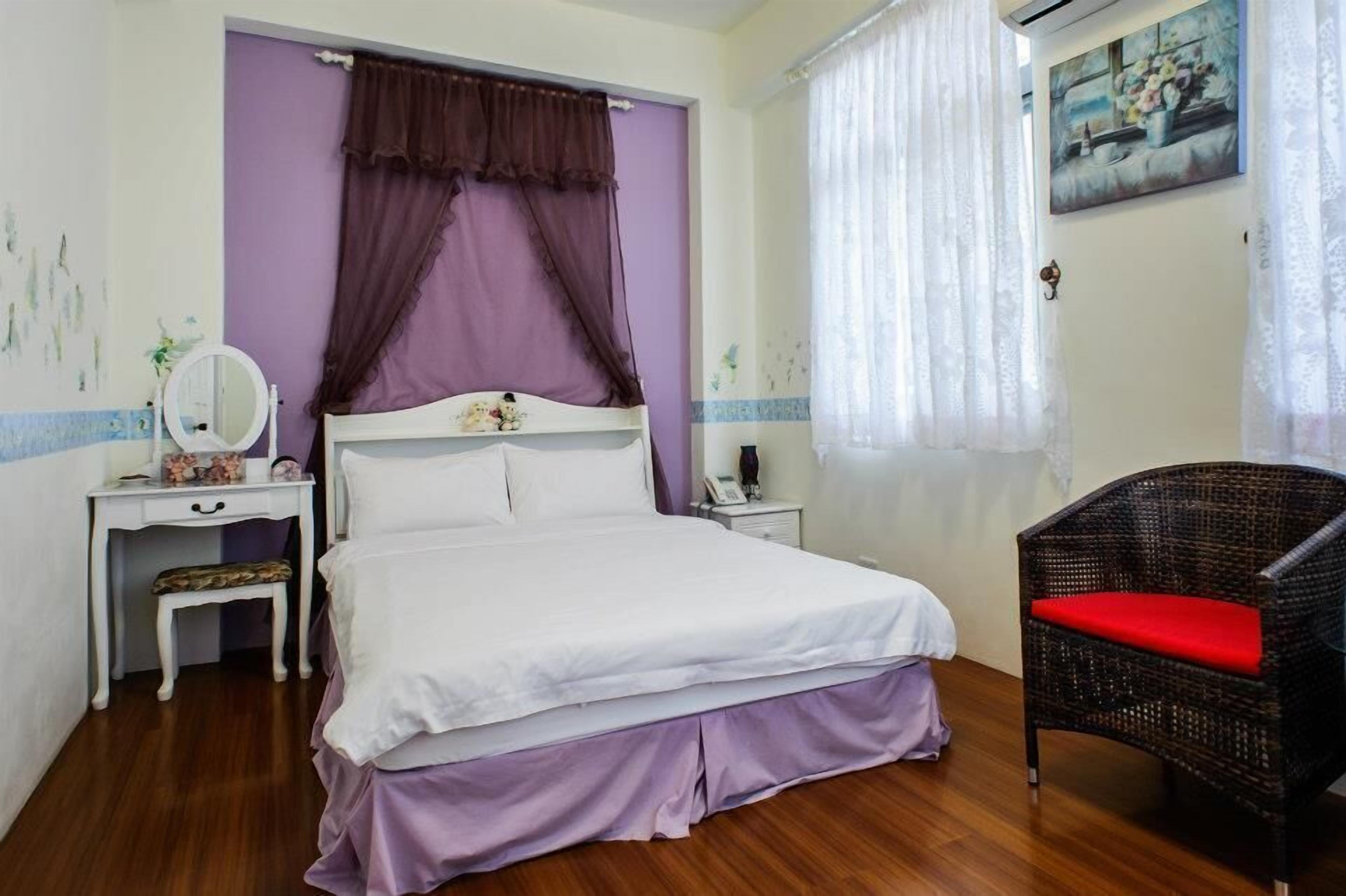 Une chambre à coucher avec un lit, une table de chevet, une chaise, une fenêtre avec des rideaux et un tableau au mur.