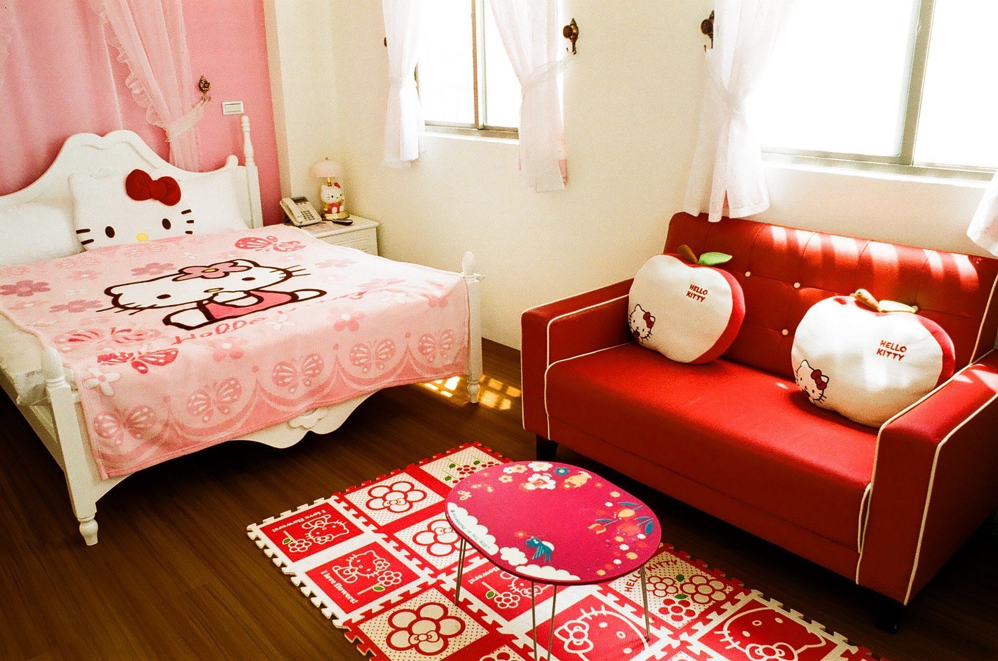 A bedroom with a pink Hello Kitty bedspread, a red sofa with Hello Kitty pillows, a small bedside table with a lamp, and a patterned rug.