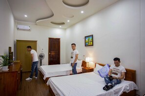 A hotel room with two beds, a wooden wardrobe, a desk with a plant, a standing fan, and a wall-mounted air conditioner.