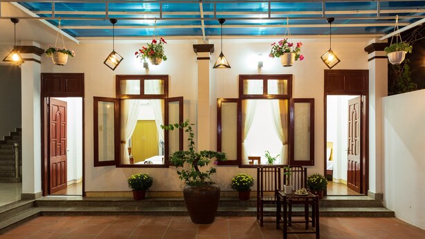 A spacious entrance with a tiled floor, large windows with wooden frames, hanging plants, and a seating area with a chair and table.