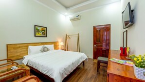 A hotel room with a wooden bed, a bedside table, a chair, a desk, a TV, and a door.