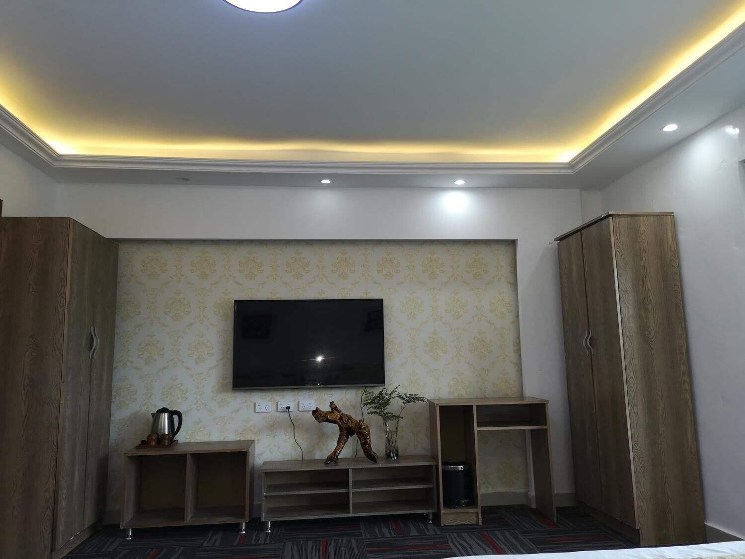 A hotel room with a flat-screen TV mounted on the wall, wooden furniture, and a patterned wallpaper.
