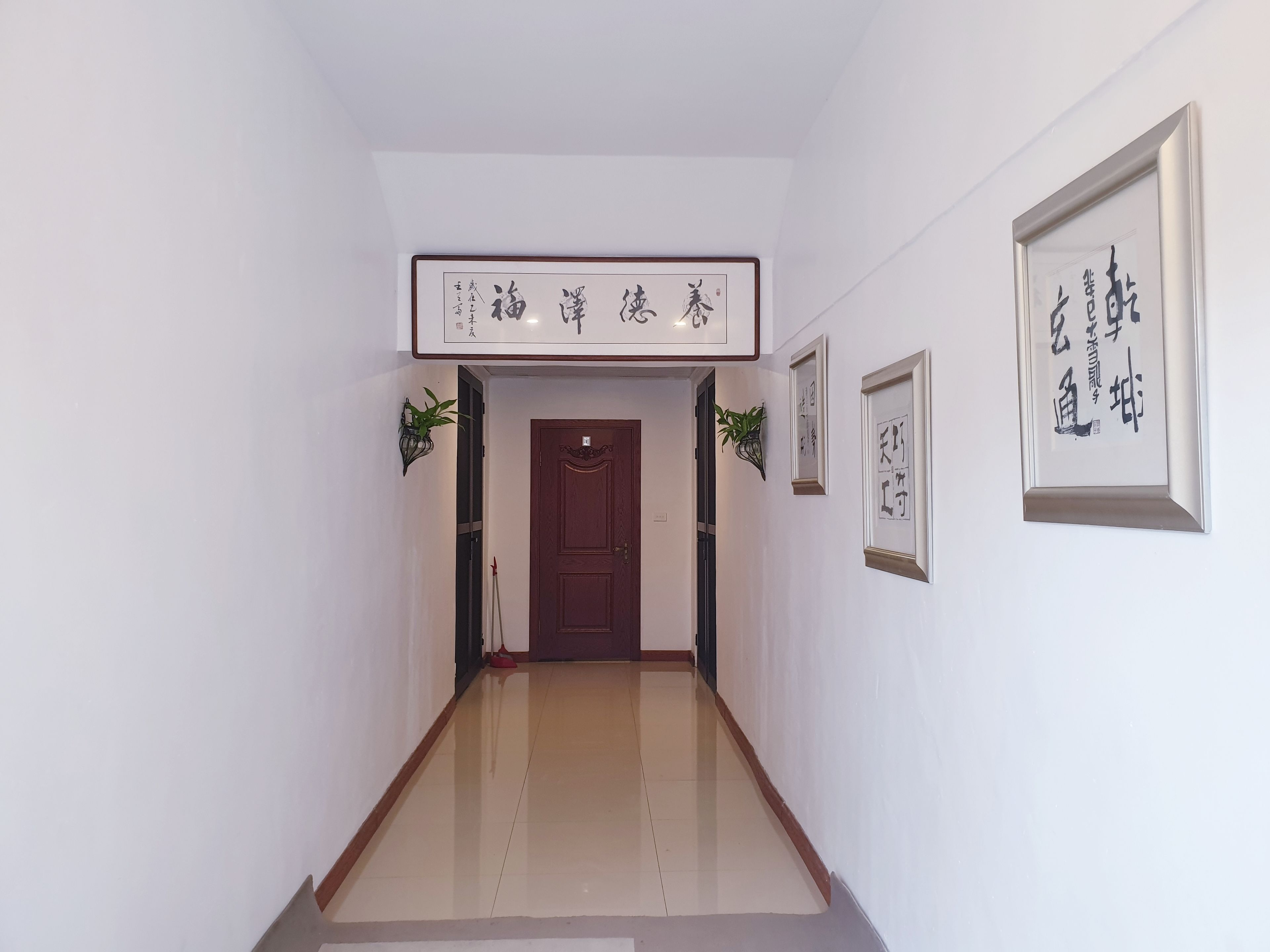 A hallway with a wooden door, framed calligraphy art, and potted plants.
