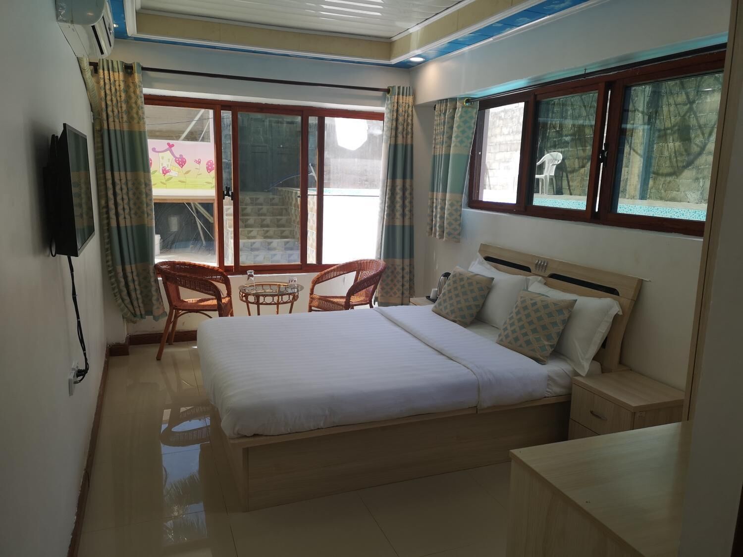 Une chambre à coucher comprenant un lit, des tables de chevet, un téléviseur, un balcon avec des meubles en osier et de grandes fenêtres.