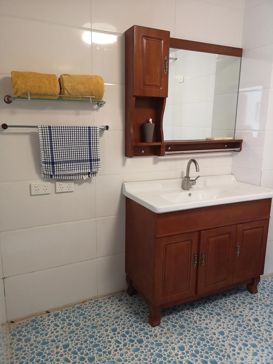 A bathroom with a wooden vanity, white sink, and a mirror above it. There are two towels hanging on a towel rack and a checkered towel on the wall.