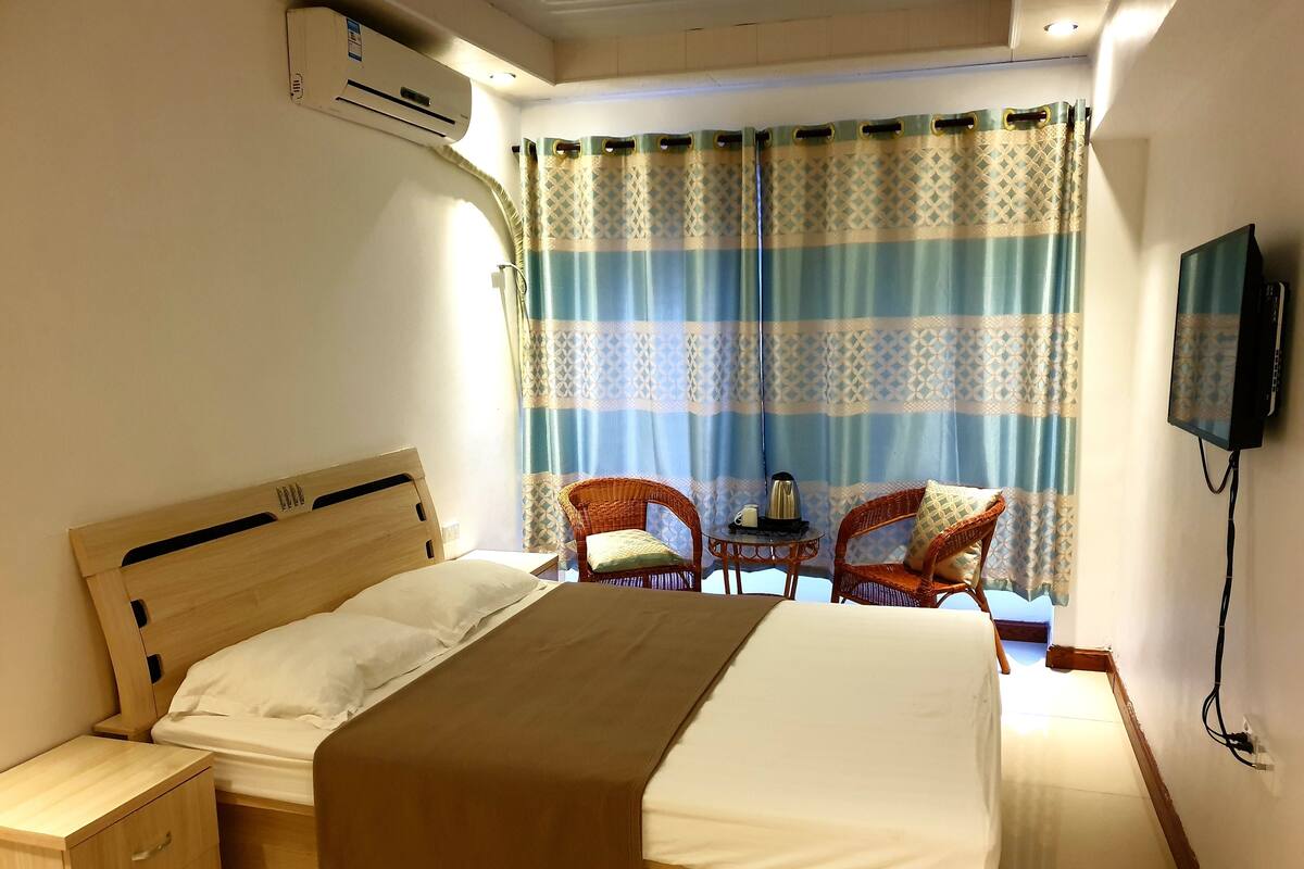 A hotel room with a wooden bed, a bedside table with drawers, a small table with a kettle and a cup, two wicker chairs, a TV mounted on the wall, and a window with patterned curtains.