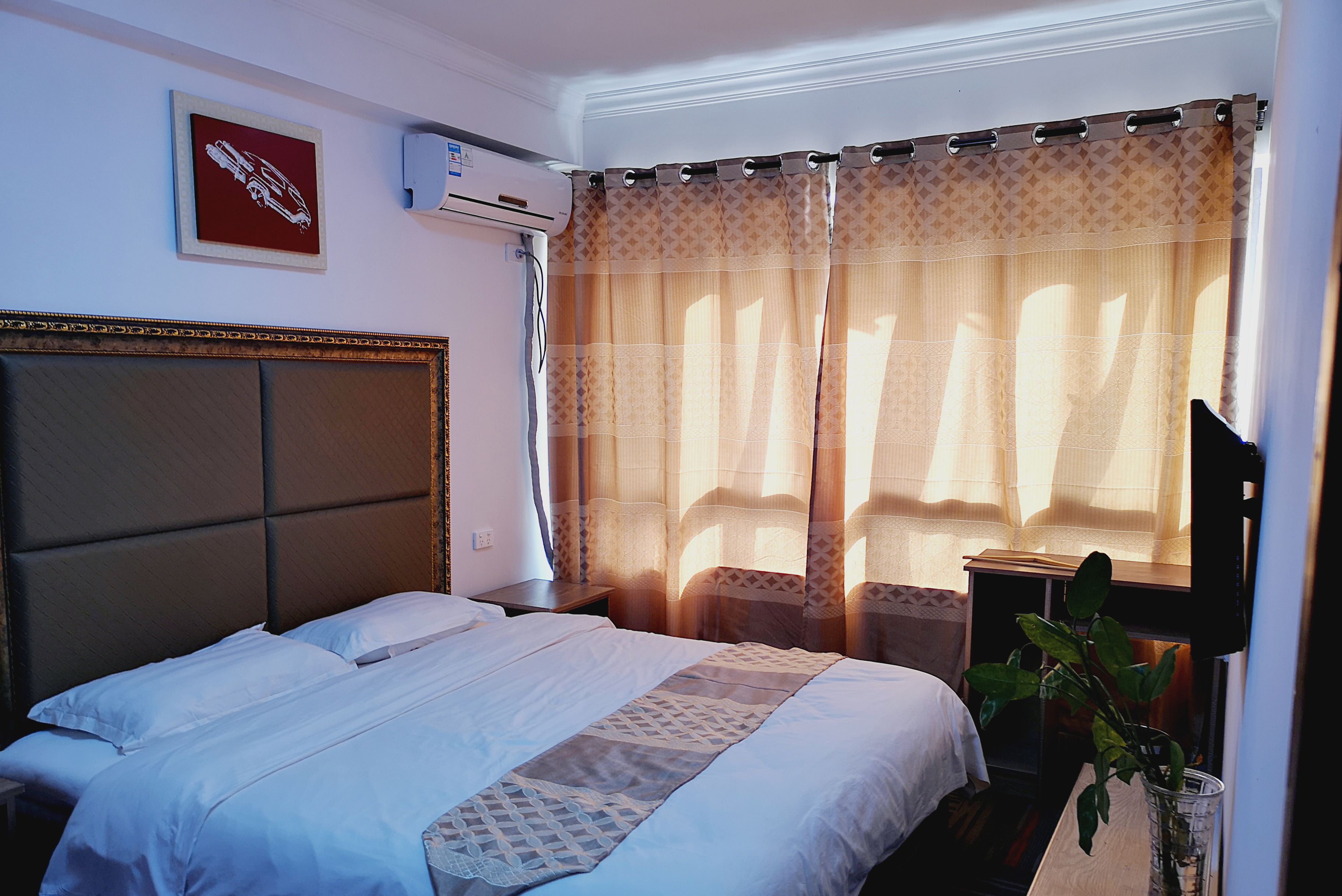 A hotel room with a bed, a bedside table, a potted plant, and a window with curtains.