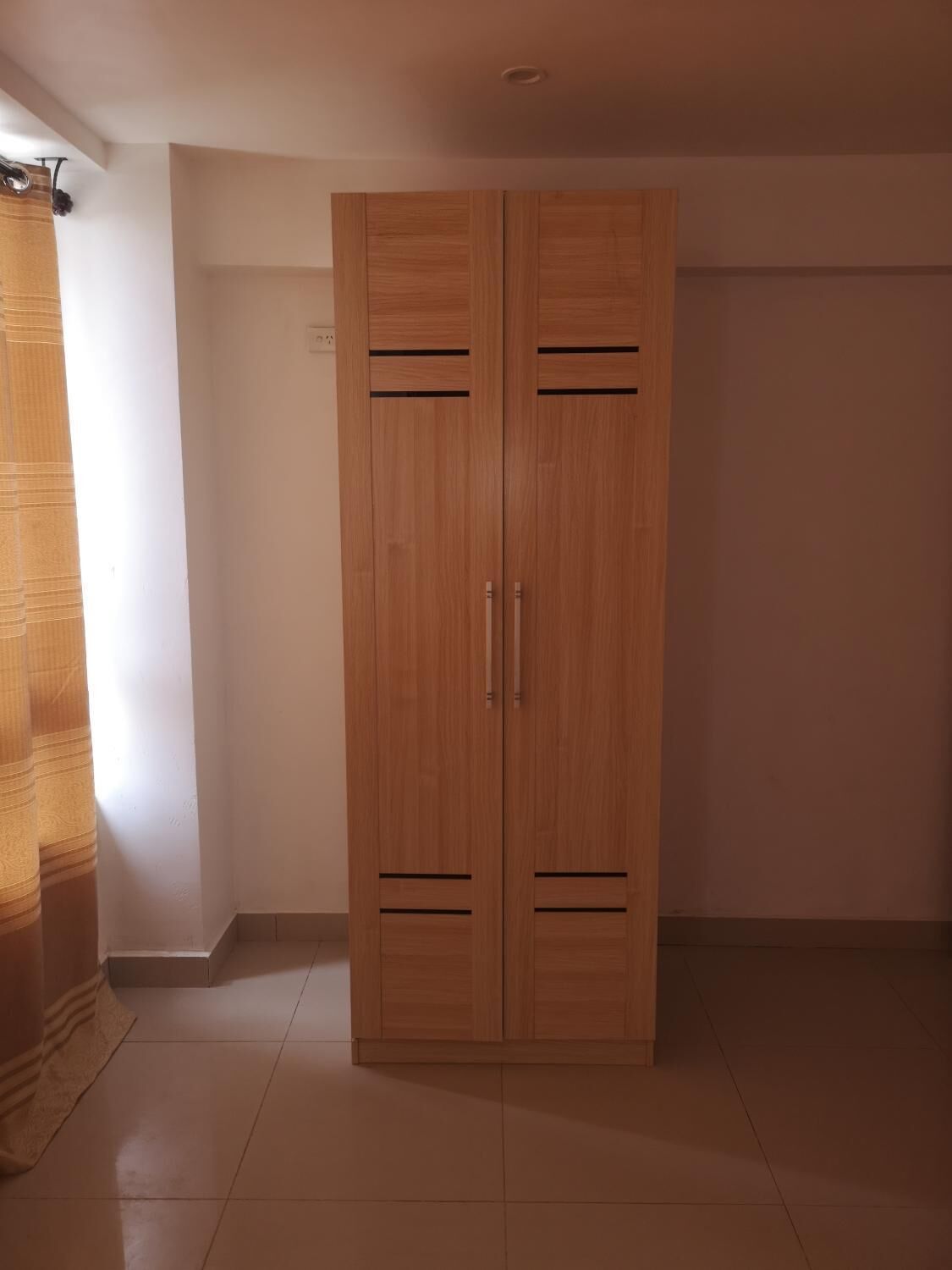 A wooden wardrobe with two doors and handles, located in a room with tiled flooring and a plain white wall.