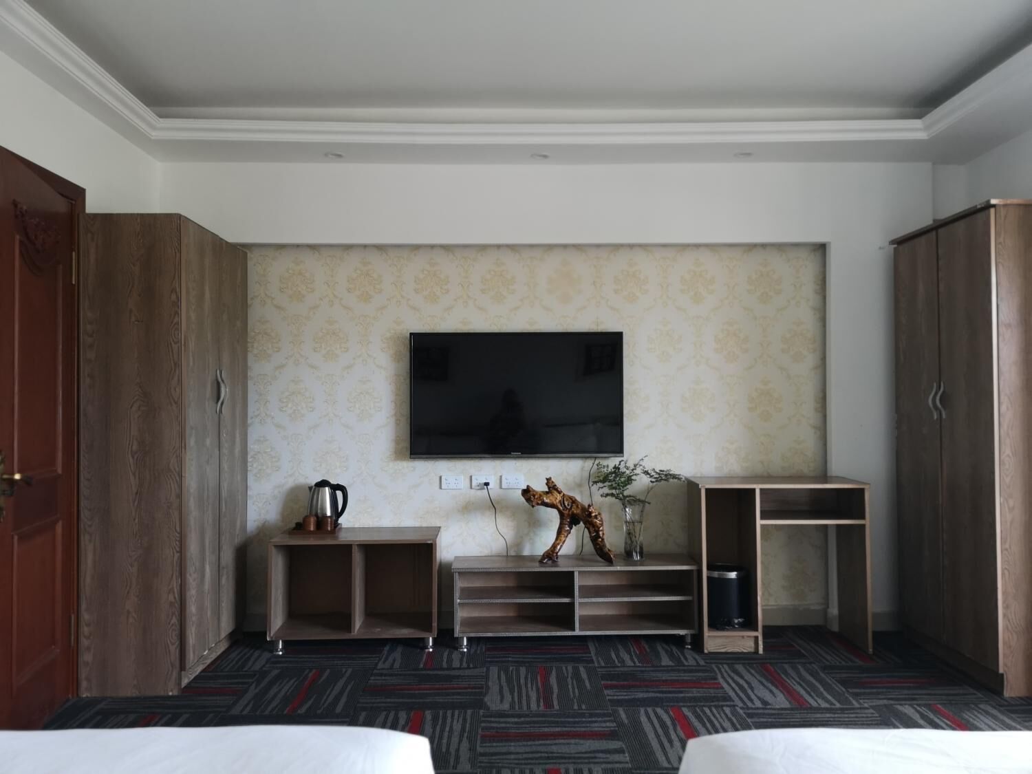 A hotel room with a large flat-screen TV mounted on the wall, a wooden cabinet, a small side table with a kettle, a decorative plant, and a bed with a striped carpet.