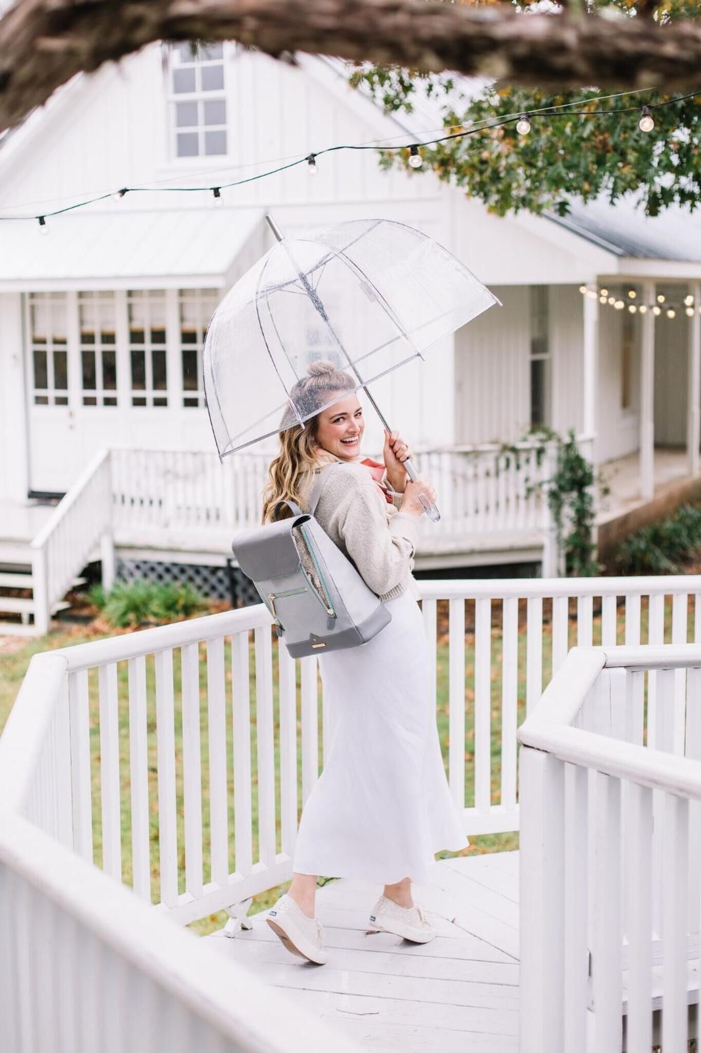 A person on a wooden deck with a white railing, holding a transparent umbrella and a gray handbag.