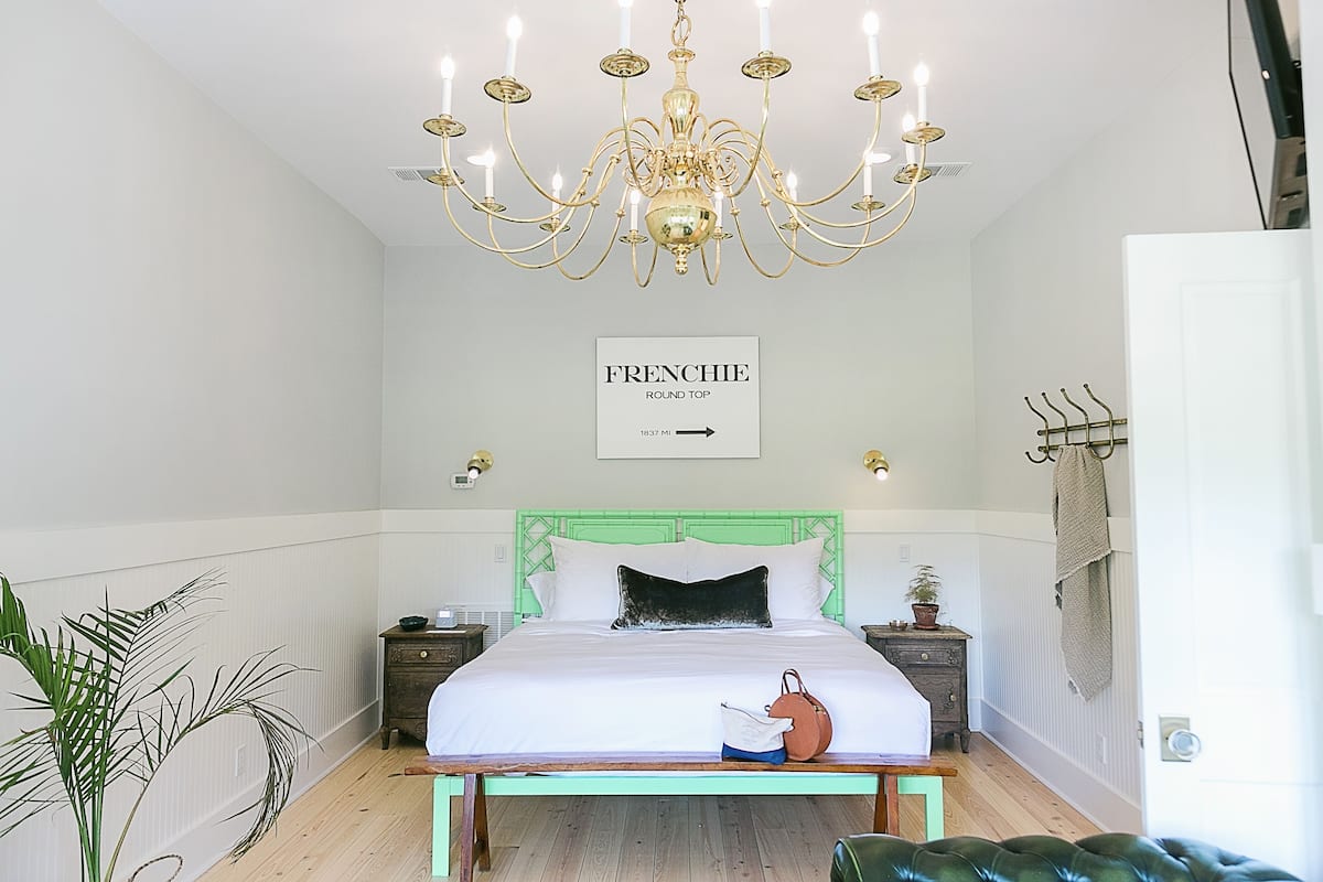 A bedroom with a green headboard, a chandelier, a bed with white bedding, and a sign that says "FRENCHIE ROUND TOP".