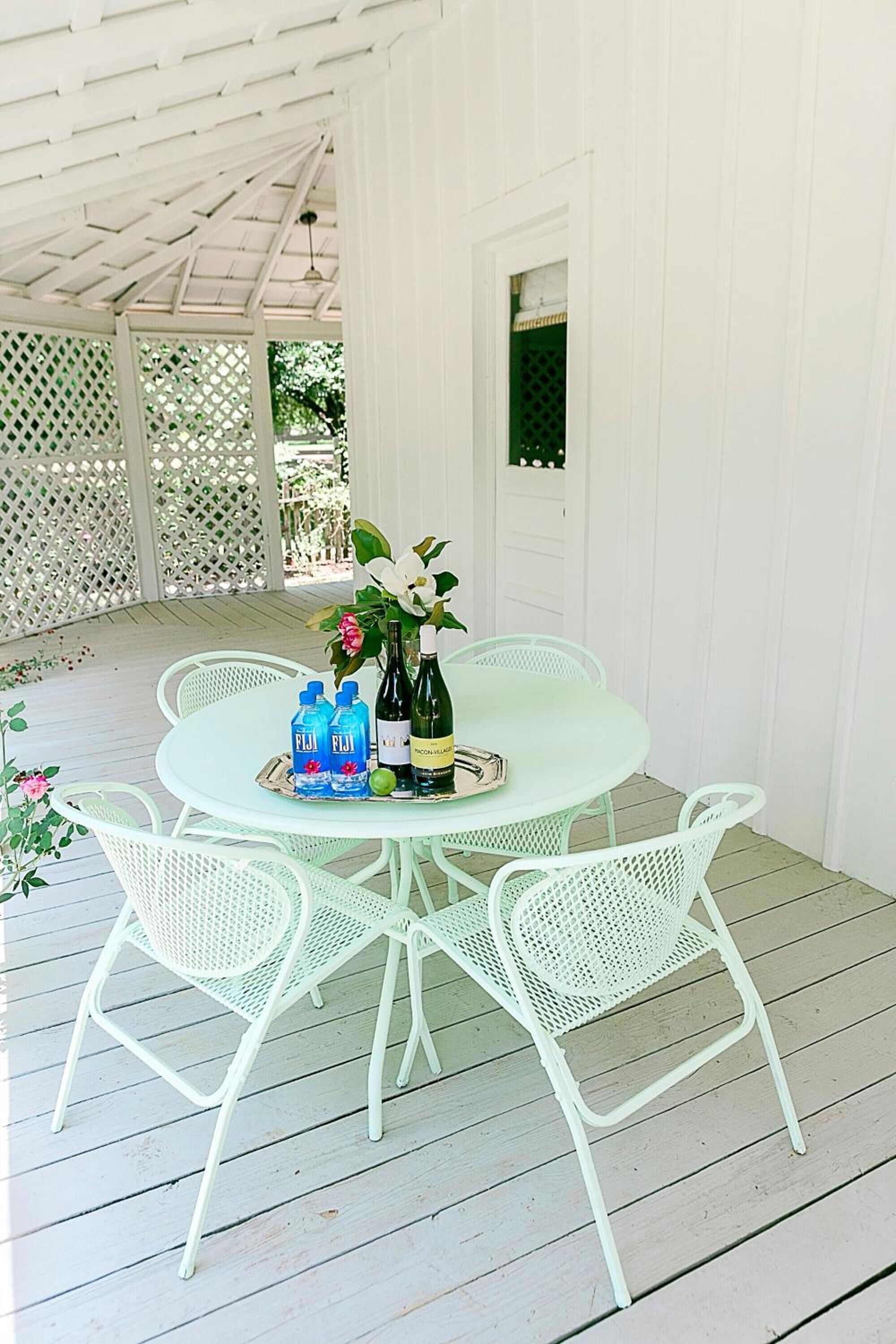A covered outdoor patio with a round table and white metal chairs, featuring a bottle of Fiji water, a bottle of wine, and a decorative flower arrangement.