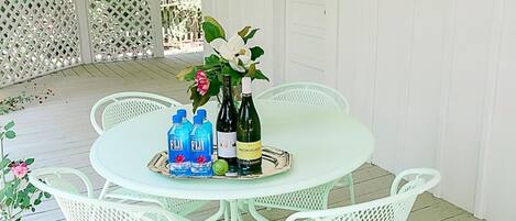 A covered outdoor patio with a round table and white metal chairs, featuring a bottle of Fiji water, a bottle of wine, and a decorative flower arrangement.